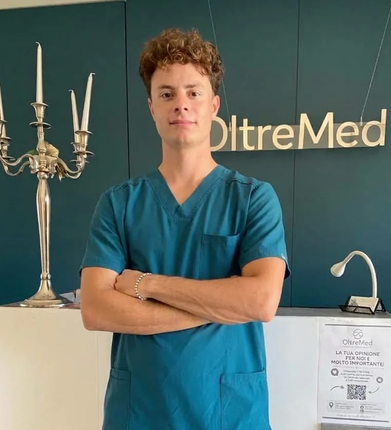 Dott. Alessandro Della Betta, in divisa verde petrolio, con le braccia incrociate davanti all’insegna OltreMed