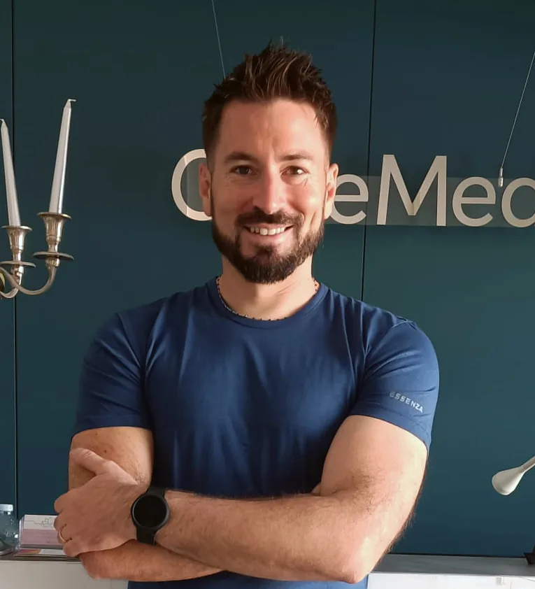 Dott. Andrea Boni, sorridente, in maglietta blu, con le braccia incrociate davanti all’insegna OltreMed
