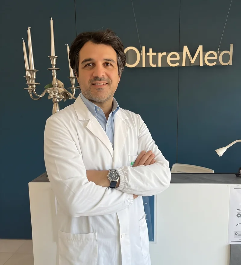 Marco Masciangelo, in camice bianco, con le braccia incrociate davanti all’insegna OltreMed