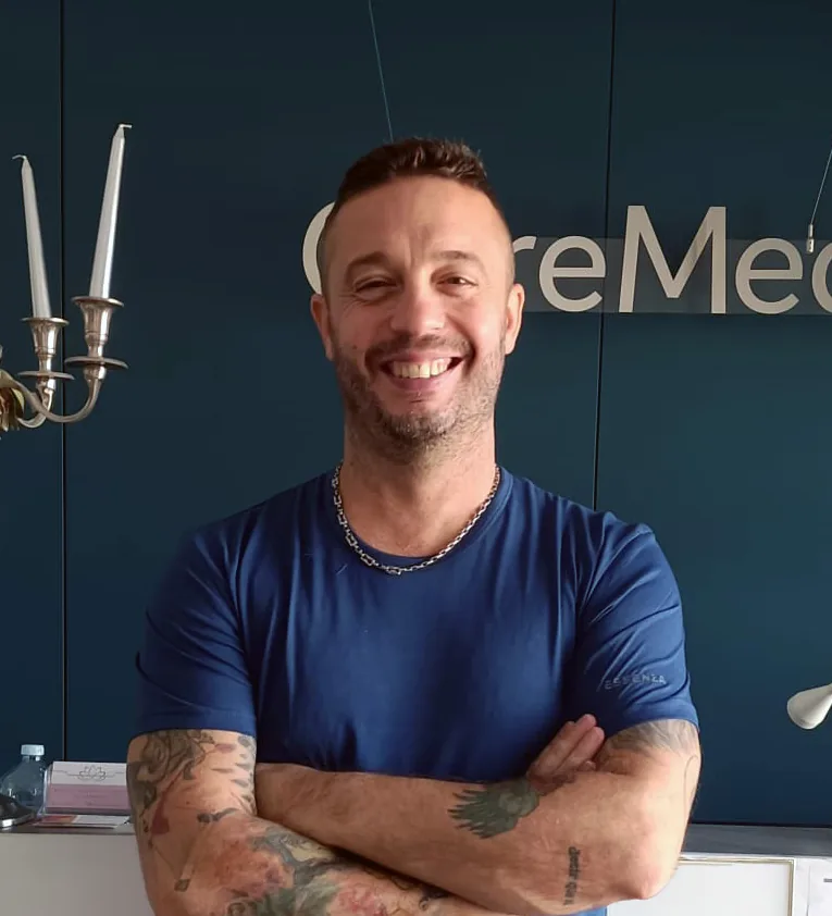 Dott. Simone Delprete, in maglietta blu, con le braccia incrociate davanti all’insegna OltreMed
